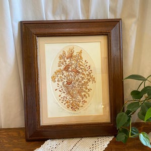 May include: A framed artwork featuring an oval-shaped botanical illustration in shades of brown, orange, and yellow. The artwork is set against a cream-colored background and framed in a dark wood frame. The artwork is signed.