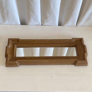 May include: A rectangular wooden mirror with a light brown frame. The mirror has a simple, classic design with decorative edges. The frame is made of wood and has a natural finish. The mirror is a decorative item.