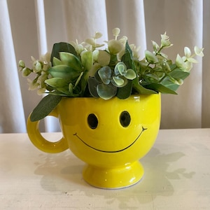 May include: A bright yellow ceramic mug featuring a black smiley face design with two circular eyes and a curved mouth. The mug is filled with a cluster of artificial green eucalyptus leaves and small white flowers, creating a decorative arrangement.