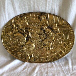 May include: A brass wall plaque depicting a scene of people gathered around a table with food and drink. The plaque is oval shaped and has a raised design.