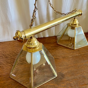 May include: A brass and glass two-light pendant light fixture with a chain hanging from the ceiling. The fixture has two square glass shades with gold trim and white frosted light bulbs.