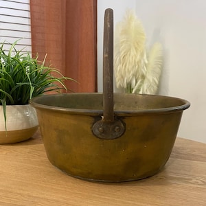 May include: A vintage brass cauldron with a single, arched handle. The cauldron has a wide, rounded bowl and a dark, aged patina. The handle is attached with rivets. The cauldron sits on a wooden surface, with a plant and window in the background.