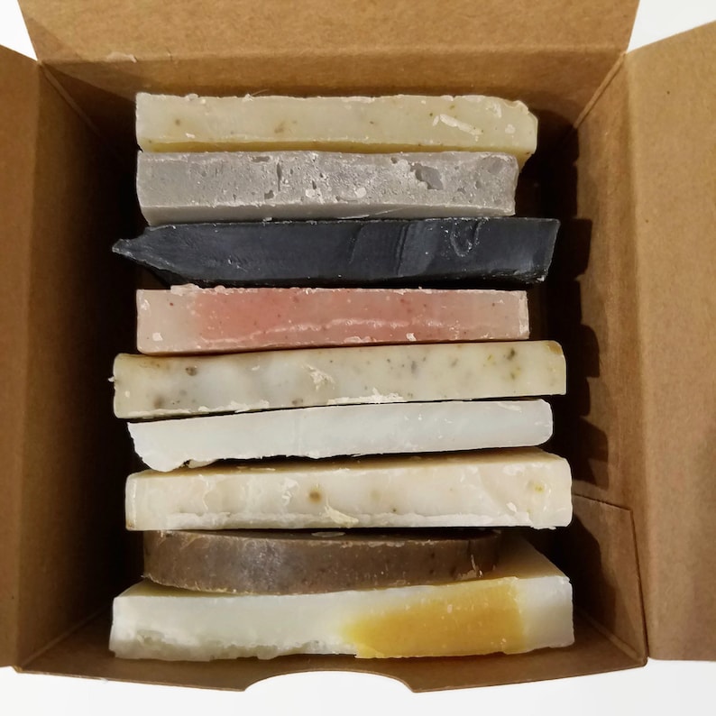 May include: A box filled with a stack of ten different colored and scented bars of soap. The soaps are arranged in a neat stack, with the top bar being a light yellow color and the bottom bar being a dark brown color.