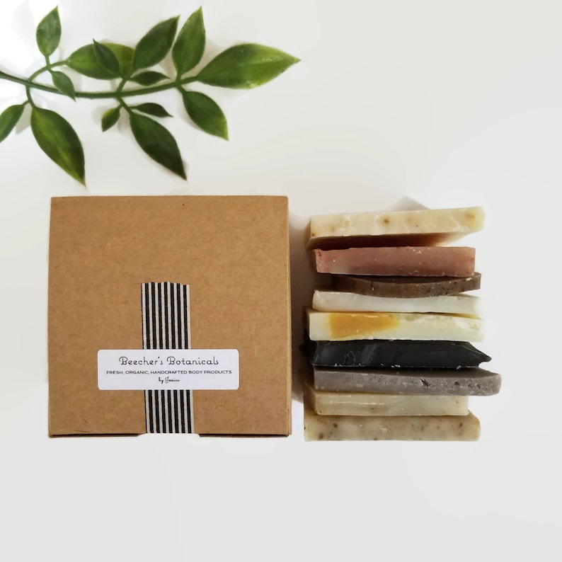 May include: A brown cardboard box with a white label that reads "Beecher's Botanicals Fresh, Organic, Handcrafted Body Products by Jamie" and a black and white striped ribbon. The box is next to a stack of eight different colored handmade soap bars.