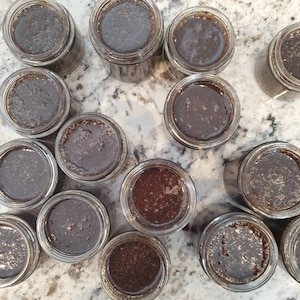 May include: A collection of 14 small glass jars filled with a dark brown liquid. The jars are arranged in a semi-circle on a white countertop with a speckled pattern.