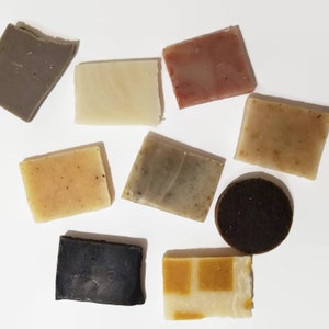 May include: Assortment of eight handmade soap bars in various colors and patterns, including gray, white, pink, brown, and beige. The soaps are arranged on a white surface.