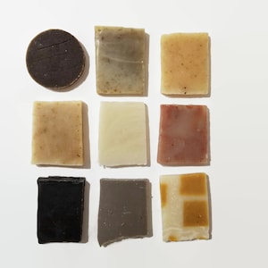 May include: Assortment of nine bars of soap in various colors and patterns, including brown, green, beige, white, pink, black, and gray. The soap bars are arranged in a grid pattern on a white surface.