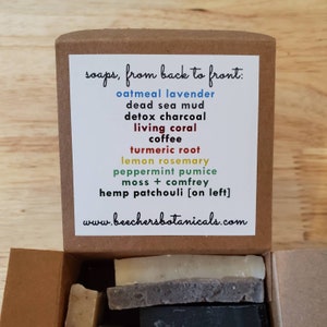 May include: A brown cardboard box with a white label listing different soap scents. The label reads "soaps, from back to front:" followed by a list of soap scents in different colors. The box is open and contains several bars of soap.