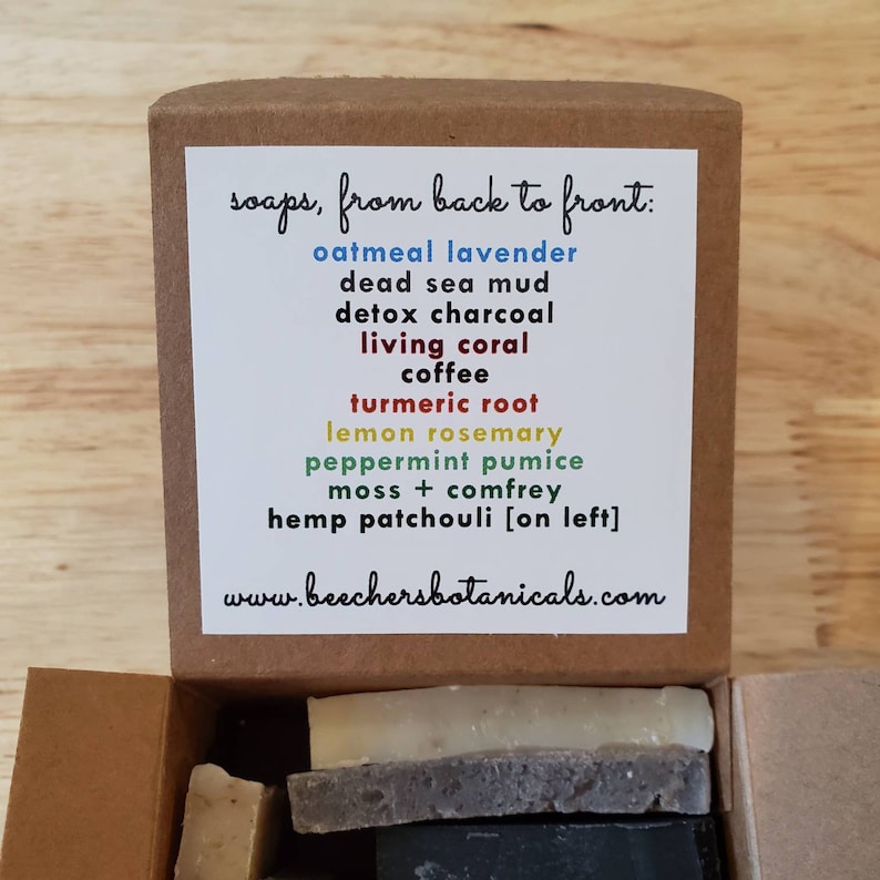 May include: A brown cardboard box with a white label listing different soap scents. The label reads "soaps, from back to front:" followed by a list of soap scents in different colors. The box is open and contains several bars of soap.