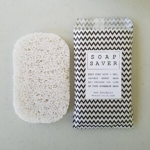 May include: A white soap saver made of natural loofah sponge. The soap saver is packaged in a black and white chevron patterned paper bag with the text "SOAP SAVER" and instructions to keep soap high and dry to prevent mushy bars and prolong the life of homemade bars. The bag also includes the website address "www.BeechersBotanicals.com".