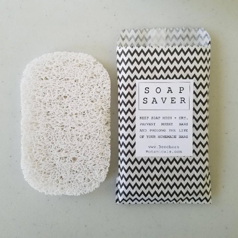 May include: A white soap saver made of natural loofah sponge. The soap saver is packaged in a black and white chevron patterned paper bag with the text "SOAP SAVER" and instructions to keep soap high and dry to prevent mushy bars and prolong the life of homemade bars. The bag also includes the website address "www.BeechersBotanicals.com".