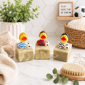 May include: Three bars of soap with soccer-themed rubber duck toppers. Each duck has a different jersey design and holds a soccer ball. The soap bars are light green and sit on a white surface. A sign reads "PLAY HAVE FUN BE KIND".