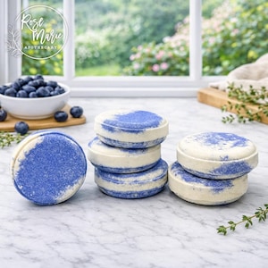 May include: A collection of round, white and blue bath bombs. The bath bombs have a layered design with a blue, speckled top. A bowl of fresh blueberries sits in the background, along with the Rose Marie Apothecary logo.