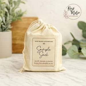 May include: A cream-colored sample sack with a drawstring closure. The sack features a rectangular label with the text "Sample Sack" and "Sniff, Sample, & Smile! Enjoy free samples on us." The Rose Marie Apothecary logo is in the upper right corner.