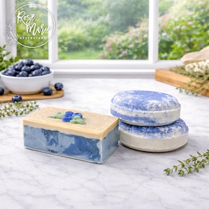 May include: A still life featuring handmade soaps and bath bombs. A rectangular soap bar with a blueberry design sits next to two round bath bombs. The soaps are blue, white, and tan. A bowl of blueberries and sprigs of thyme are also present.