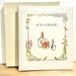 Könnte beinhalten: Ein cremefarbenes Sammelalbum mit vertikalem Nadelstreifenmuster und einer zentralen Illustration von Peter Hase und seiner Familie, umgeben von einer Blumenbordüre. Das Wort SCRAPBOOK ist in blauer Schrift über den Hasen abgedruckt.