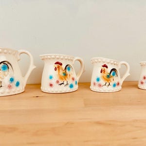 May include: A set of four cream-colored ceramic pitchers. Each pitcher features a hand-painted rooster design with red, yellow, blue, and black details. The pitchers vary in size, with the largest measuring approximately 7 inches tall.