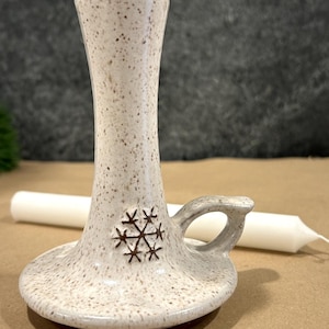May include: White ceramic candle holder with a snowflake design and a brown handle. The holder has a speckled glaze.