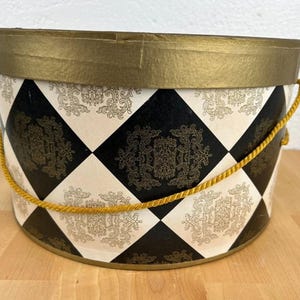 Peut inclure: Une boîte à chapeau ovale décorative avec un bord et une poignée dorés. La boîte présente un motif arlequin noir et blanc avec des motifs de filigrane dorés. La boîte est en carton.