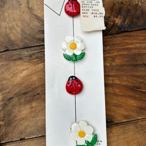 May include: A vertical white display card featuring four decorative buttons: two red ladybugs with black spots and two white flowers with yellow centers and green leaves. The card includes a price tag and the text Casey Coleman and Made in U.S.A.