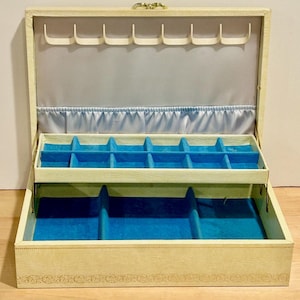May include: Open vintage jewelry box with a cream-colored exterior and a gold clasp. The interior features a light blue lining and multiple compartments with blue velvet lining for storing jewelry.