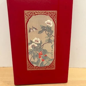 May include: A rectangular red book with a central vertical panel featuring a beige background, white flowers, green leaves, red berries, and a butterfly. The panel is framed by a gold geometric border and includes black Asian characters.