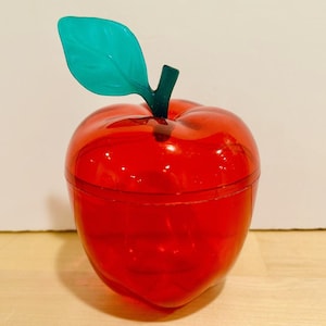 May include: A translucent red apple-shaped container with a green leaf and stem. The container is made of a shiny material and has a lid. The apple-shaped container is a decorative item.