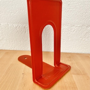 May include: A bright red metal bookend featuring a tall, rectangular upright panel with a central arched cutout and a flat base extending backward with a single circular hole. The surface has a smooth finish with embossed text near the base.