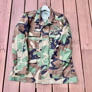 May include: A camouflage field jacket featuring a woodland pattern of brown, green, and black. The jacket includes four front pockets, a collar, and buttoned cuffs. Patches on the chest display the text U.S. ARMY along with military insignia.