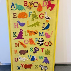 May include: A yellow-framed wall hanging featuring a yellow fabric background with the alphabet from A to Z embroidered in various colors. Each letter is accompanied by a colorful yarn-stitched animal or creature illustration representing that letter.