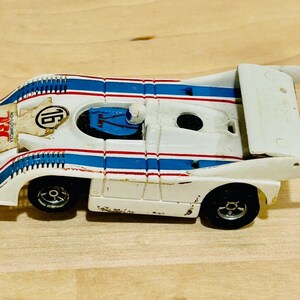 May include: A vintage white toy race car with blue and red stripes, featuring the number 16 and sponsor logos. The car has black wheels and a rear wing, showing signs of wear.