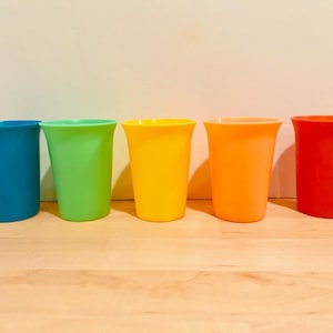 May include: Five brightly coloured plastic cups. The cups are arranged in a row and are in shades of blue, green, yellow, orange, and red. They have a flared rim and a smooth finish. They are on a light-coloured wooden surface.
