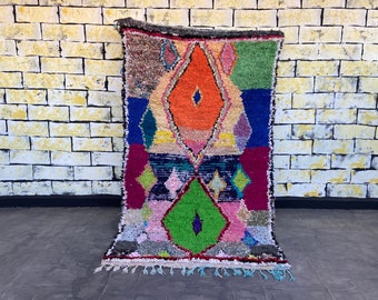 May include: A colorful Moroccan rug with a geometric pattern in shades of red, blue, green, orange, and pink. The rug has a fringe border and is hanging on a brick wall.
