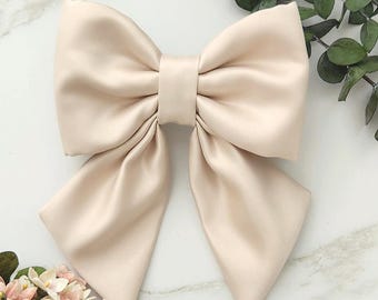 May include: A large, champagne-colored satin bow featuring two wide, folded upper loops and two long, pointed tails. The fabric has a smooth, lustrous finish with a central knot detail that secures the structure of the bow.