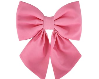 May include: A large, solid pink hair bow featuring two wide top loops and two long, pointed bottom tails. The bow is constructed from a smooth, matte fabric and is centered with a narrow, horizontal fabric band that secures the structure together.