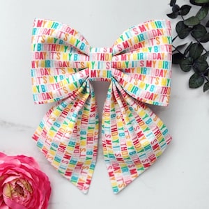 Peut inclure: Un nœud pour cheveux en tissu arborant un motif répétitif et coloré avec le texte IT'S MY BIRTHDAY dans des teintes vives. Le nœud se compose de deux grandes boucles supérieures et de deux longs pans pointus tombant vers le bas.