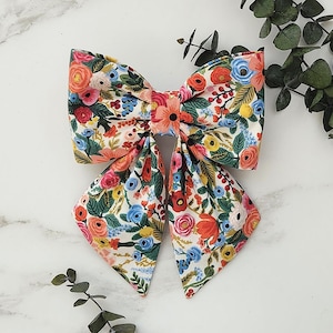 Peut inclure: Un nœud pour cheveux en tissu arborant un motif floral vibrant sur fond crème. Le design présente diverses fleurs dans des tons de rouge, orange, rose, jaune et bleu, agrémentées de feuilles et de tiges vertes, noué en forme de nœud classique.
