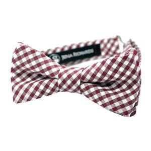 May include: A burgundy and white checkered bow tie. The bow tie is made of fabric and has a classic gingham pattern. The brand name "Jena Richards" is printed on a black label.