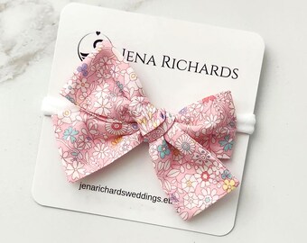 May include: A pink fabric bow hair accessory with a multi-colored floral pattern attached to a white display card featuring the text JENA RICHARDS and jenarichardsweddings.eu. The bow is secured to a thin white elastic headband.