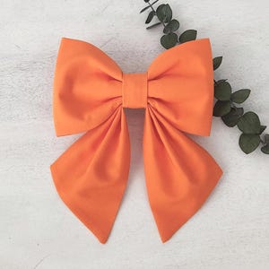 May include: A large, solid orange fabric bow featuring two wide top loops and two long, pointed bottom tails. The bow is constructed with a central fabric knot and displays a smooth, matte texture throughout its entire surface area.
