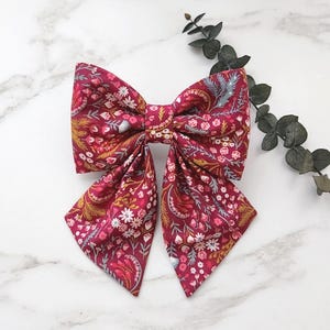 Könnte beinhalten: Eine große, bordeauxfarbene Stoffschleife mit einem Blumenmuster mit rosa, gelben und grauen Akzenten. Die Schleife ist zentriert und hat lange, fließende Enden. Der Hintergrund ist eine weiße Marmoroberfläche.