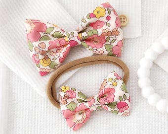 May include: Two fabric hair bows featuring a floral pattern with pink, yellow, and green colors on a cream background. One bow is attached to a thin, tan elastic headband, while the other is a standalone bow accessory with a central knot detail.