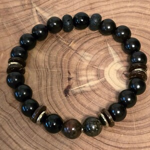 May include: A beaded bracelet featuring large, glossy black beads interspersed with smaller, textured dark beads and decorative brown and gold accents. The bracelet is displayed on a wooden surface, showcasing its natural tones and textures.