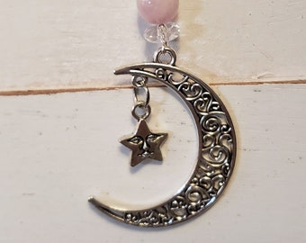 May include: A celestial-themed necklace featuring a silver crescent moon pendant with a star charm. The necklace includes a pink gemstone and clear crystal beads, suspended from a black cord. The moon pendant has an ornate, swirling design.