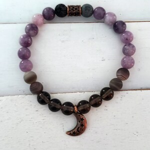 May include: A purple and grey gemstone bracelet with a crescent moon charm. The bracelet has a dark grey lava bead and a copper spacer bead.