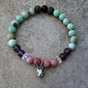 May include: A green, purple and pink beaded bracelet with a silver rabbit charm. The bracelet is made with various stones, including amethyst, rose quartz and green aventurine.