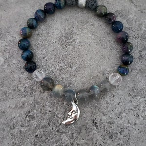 May include: A blue and grey beaded bracelet with a silver crescent moon charm. The bracelet is made with tiger's eye, labradorite, and lava rock beads.