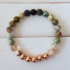 May include: A multi-colored gemstone bracelet with a gradient of green, brown, and white stones. The bracelet has a copper-colored metal accent.