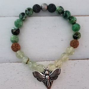May include: A green and brown beaded bracelet with a silver moth charm. The bracelet is made with various types of beads, including green agate, brown wood, and black lava rock.