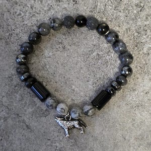 May include: A black and grey beaded bracelet with a silver wolf charm. The bracelet is made with various types of stones, including black onyx, hematite, and grey agate.